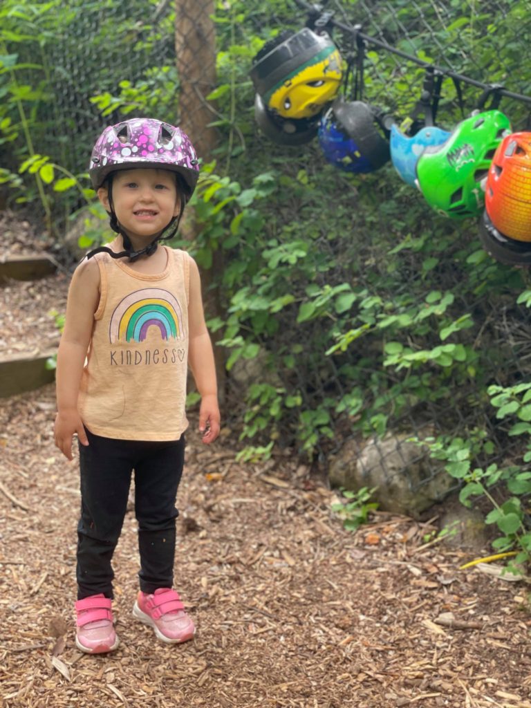 Little Girl wearing a helmet and smiling in front of a string of helmets at Oyama Zipline Adventure Park